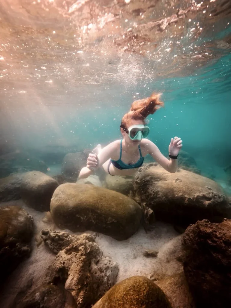 Schnorcheln auf Koh Tao: 7 Spots, die sich wirklich lohnen 10 Junge Frau schnorchelt über runde Felsen und sandigen Meeresgrund bei Shark Bay, während Sonnenstrahlen durchs Wasser brechen