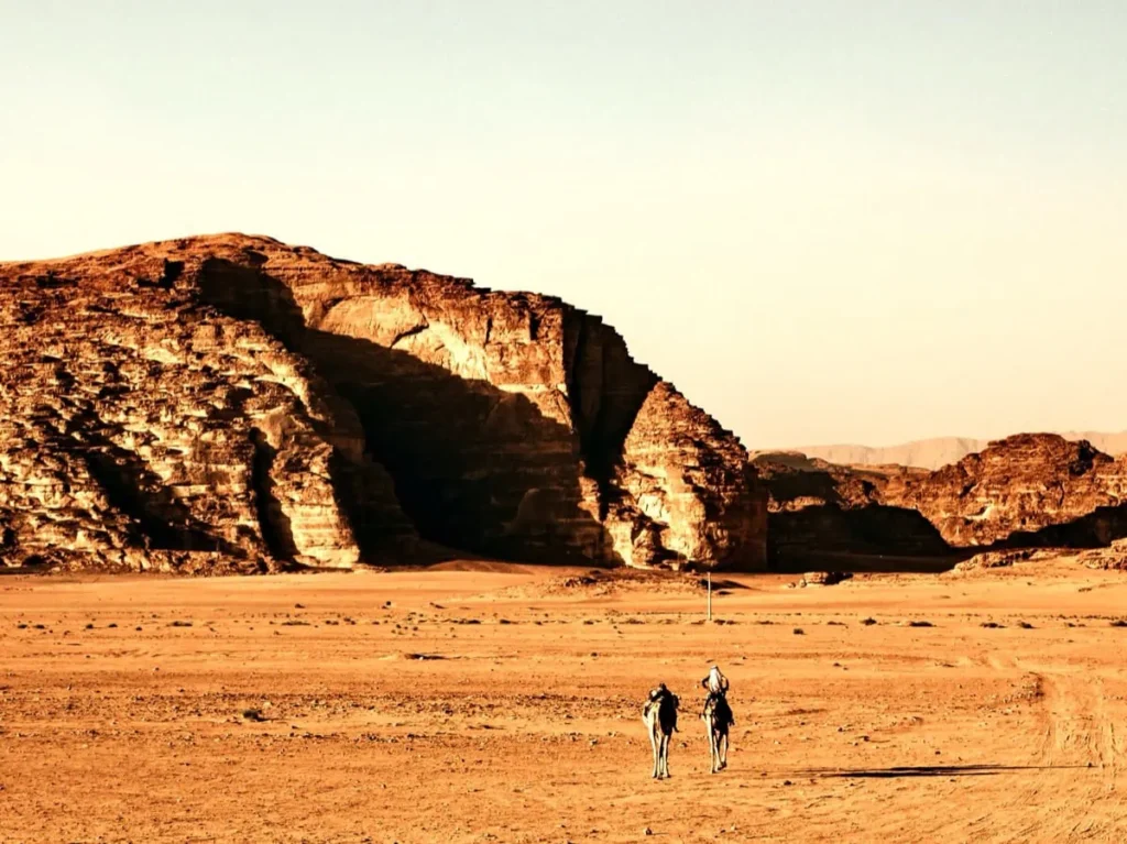 Zwei Personen reiten auf Kamelen durch die goldene Wüste von Wadi Rum, während gewaltige Sandsteinfelsen lange Schatten in der Abendsonne werfen.