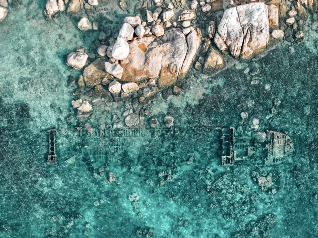 Schnorcheln auf Koh Tao: 7 Spots, die sich wirklich lohnen 1 Drohnenaufnahme eines flachen Schiffswracks bei Mae Haad auf Koh Tao, umgeben von türkisfarbenem Wasser und großen Felsen – ein besonderer Spot zum Schnorcheln auf Koh Tao.
