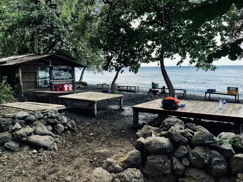 USAT Liberty Wrack Tauchguide: Tulambens berühmtes Wrack 10 Zugang zum Liberty-Wrack-Tauchplatz in Tulamben, Bali, mit hölzernen Vorbereitungsplattformen unter schattigen Bäumen, einer kleinen Hütte und dem Ozean im Hintergrund.