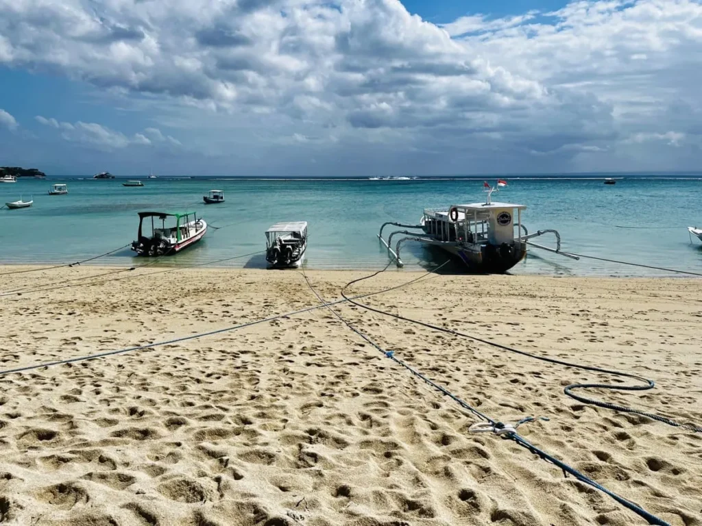 Diving Nusa Penida: The Full Guide to Mantas, Molas & Drifts 6 Three small boats anchored on the sandy beach of Nusa Penida with calm turquoise waters and cloudy skies.