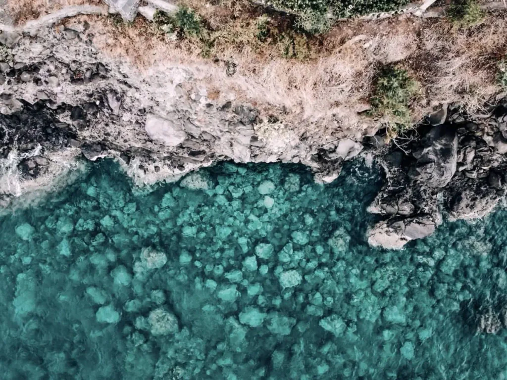 A top-down view of Nusa Penida's rugged coastline with crystal-clear turquoise water and visible underwater rocks.
