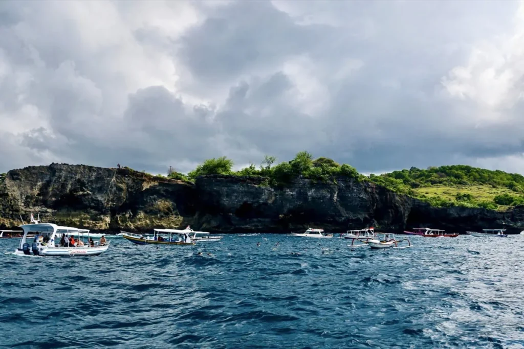 Best Snorkeling Spots in Nusa Penida + Safety Tips 2 Tour boats float near rocky cliffs in Nusa Penida while visitors snorkel in the water below.