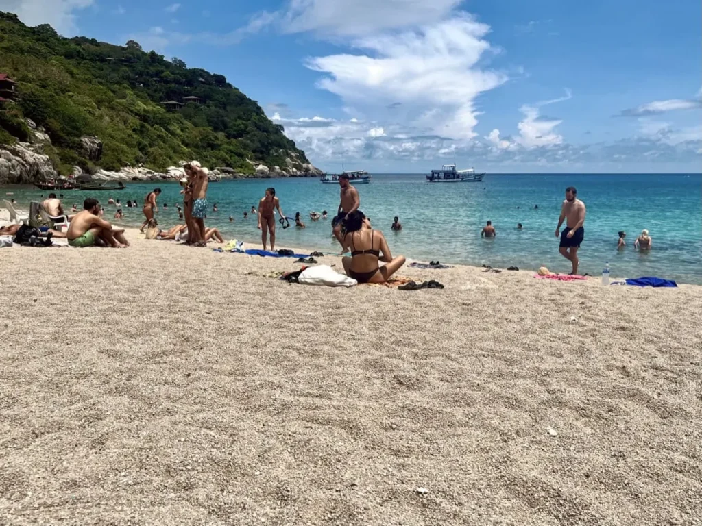 Schnorcheln auf Koh Tao: 7 Spots, die sich wirklich lohnen 5 Ein weiter Blick auf den Aow Leuk Beach zeigt Leute, die am Sandstrand entspannen und im klaren, türkisfarbenen Wasser schwimmen. Einige sitzen auf Handtüchern oder waten im Meer, während draußen Boote vor der Küste liegen, mit einem üppig grünen Hügel im Hintergrund. Das Bild vermittelt eine lebendige Strandstimmung an einem sonnigen Tag, mit ruhigem Wasser und leichten Wolken am Himmel.