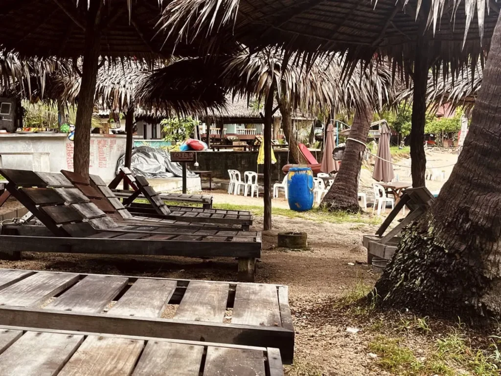 Schnorcheln auf Koh Tao: 7 Spots, die sich wirklich lohnen 4 Holzliegen stehen leer unter geflochtenen Palmenschirmen am Sandstrand von Aow Leuk, im Schatten hoher Palmen. Im Hintergrund sieht man Plastikstühle, Tische, geschlossene Schirme und einen kleinen Beachbar-Bereich, was der Szene ein ruhiges Gefühl außerhalb der Stoßzeiten gibt. Das Bild zeigt eine entspannte, leicht rustikale Strandumgebung ganz ohne Menschen.