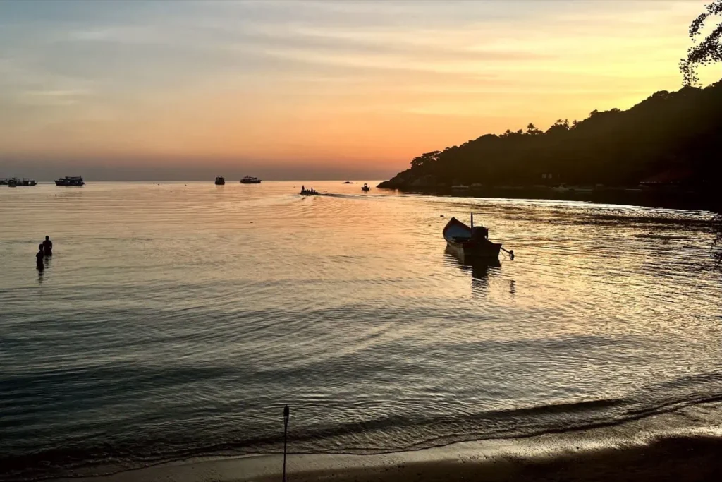 Ein dramatischer rot orangefarbener Sonnenuntergang über dem Chalok Baan Kao Beach auf Koh Tao, mit einem Holzboot, das im Sand liegt, und beleuchteten Resorts am Hang im Hintergrund. Chalok Baan Kao gehört zu den besten Gegenden, um auf Koh Tao zu wohnen, wenn du eine ruhige Atmosphäre abseits der Partybereiche suchst.