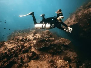 Eine Tauchlehrerin in Sidemount-Ausrüstung erkundet ein felsiges Riff auf Koh Tao. Sie trägt ein schwarzes Bandana und eine Tauchmaske und schwimmt nahe an einer dunklen Felsspalte, umgeben von kleinen Fischen. Das klare blaue Wasser und die zerklüfteten Korallenformationen schaffen eine beeindruckende Unterwasserszene.