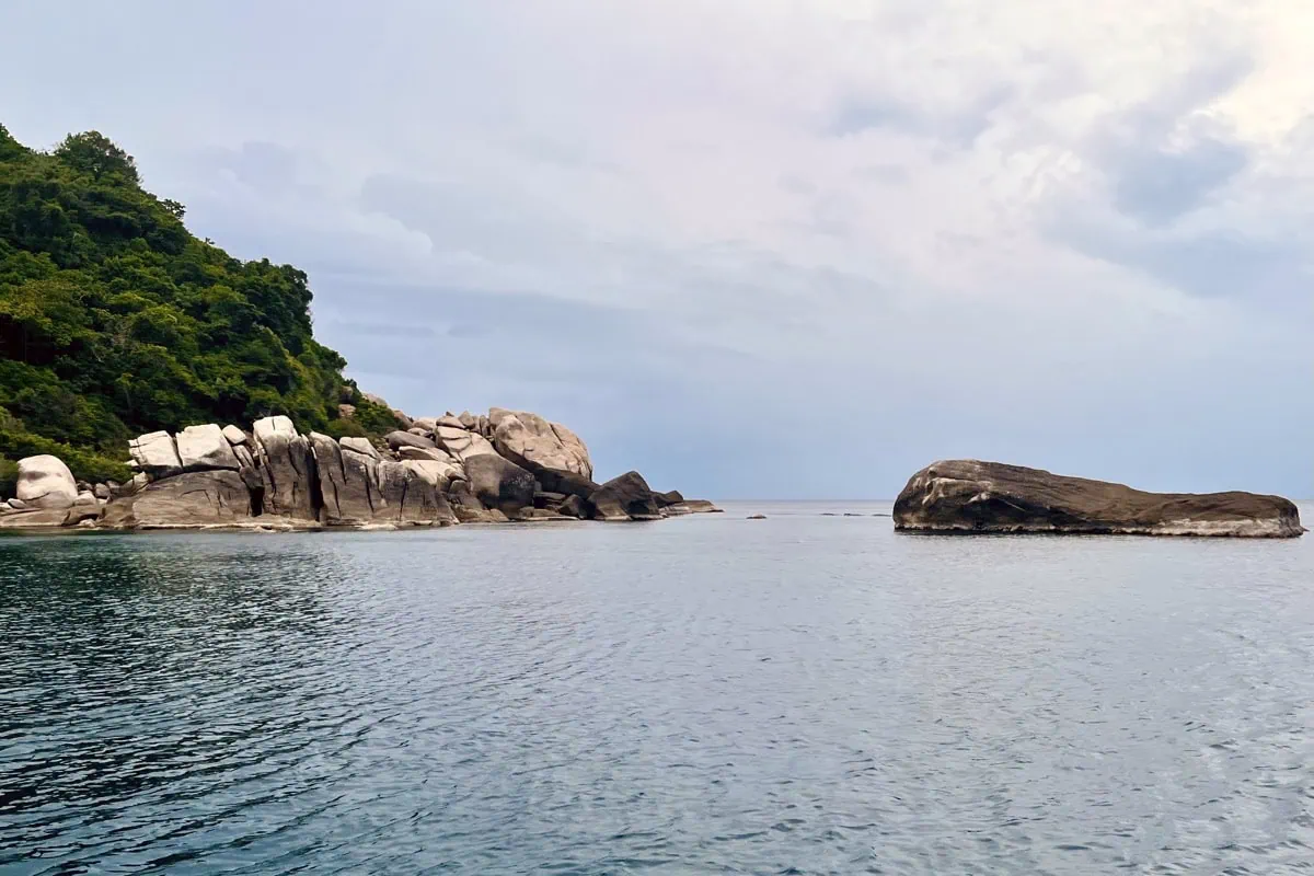 A calm view of Lighthouse Bay shows smooth blue water surrounded by large granite rocks and a forested hillside. A long low rock formation sits offshore while rounded boulders line the shoreline under a lightly clouded sky. The image emphasizes the quiet secluded feel of Lighthouse Bay and its dramatic rock formations.