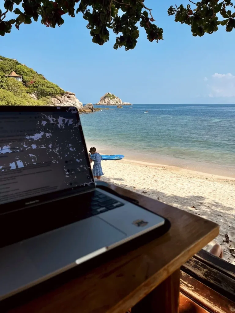 Schnorcheln auf Koh Tao: 7 Spots, die sich wirklich lohnen 7 Laptop auf einem Holztisch mit Blick auf das Meer und den Strand von Sai Daeng. Im Hintergrund liegt die geschützte Bucht mit ruhigem Wasser.