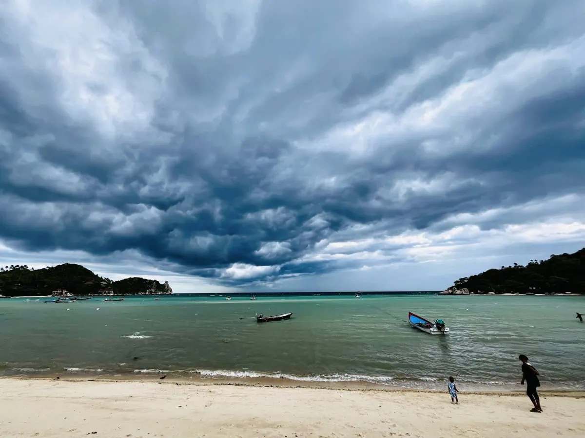 Beste Zeit zum Tauchen auf Koh Tao: Tipps von einer Tauchlehrerin 4 Dramatischer Himmel über der Chalok Bay auf Koh Tao, während dunkle Sturmwolken über dem türkisfarbenen Wasser aufziehen. Traditionelle Holzboote treiben nahe am Ufer, ein Kind und ein Erwachsener spazieren am Sandstrand entlang. Im Hintergrund rahmen grüne Hügel die tropische Kulisse
