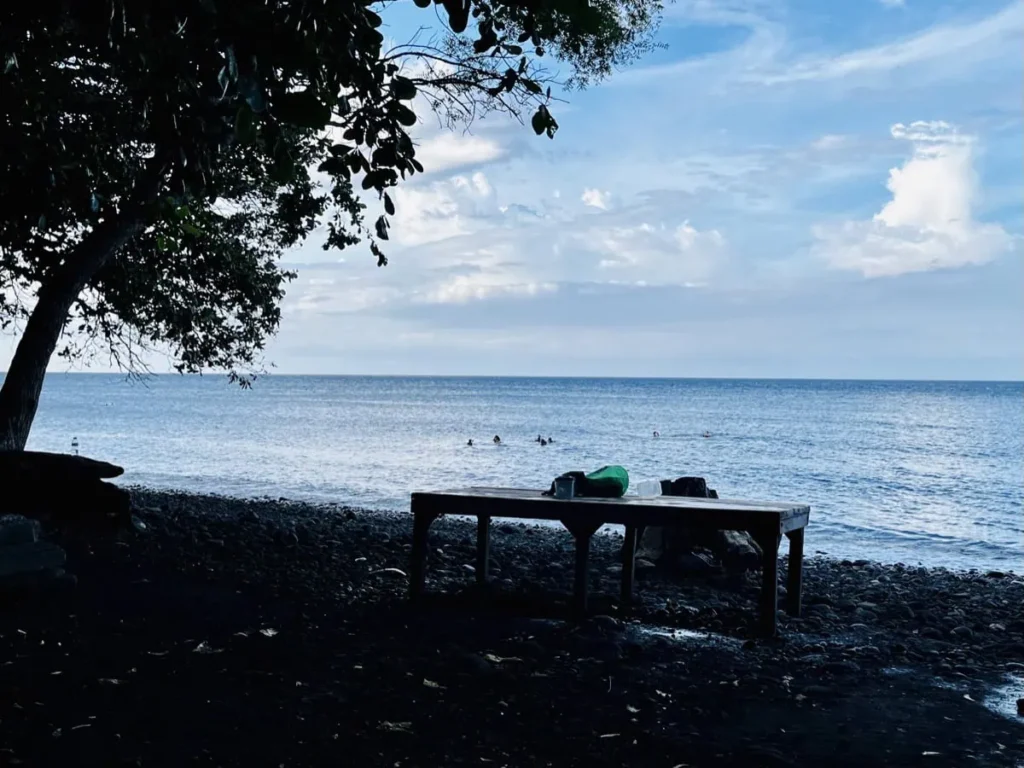 Die besten Aktivitäten in Tulamben zwischen den Tauchgängen 2 Schnorchler im ruhigen Meer vor dem dunklen Vulkanstrand von Tulamben, während am Ufer unter einem Baum ein Tisch mit Schnorchelausrüstung steht und der Blick über das offene Wasser hinausgeht.
