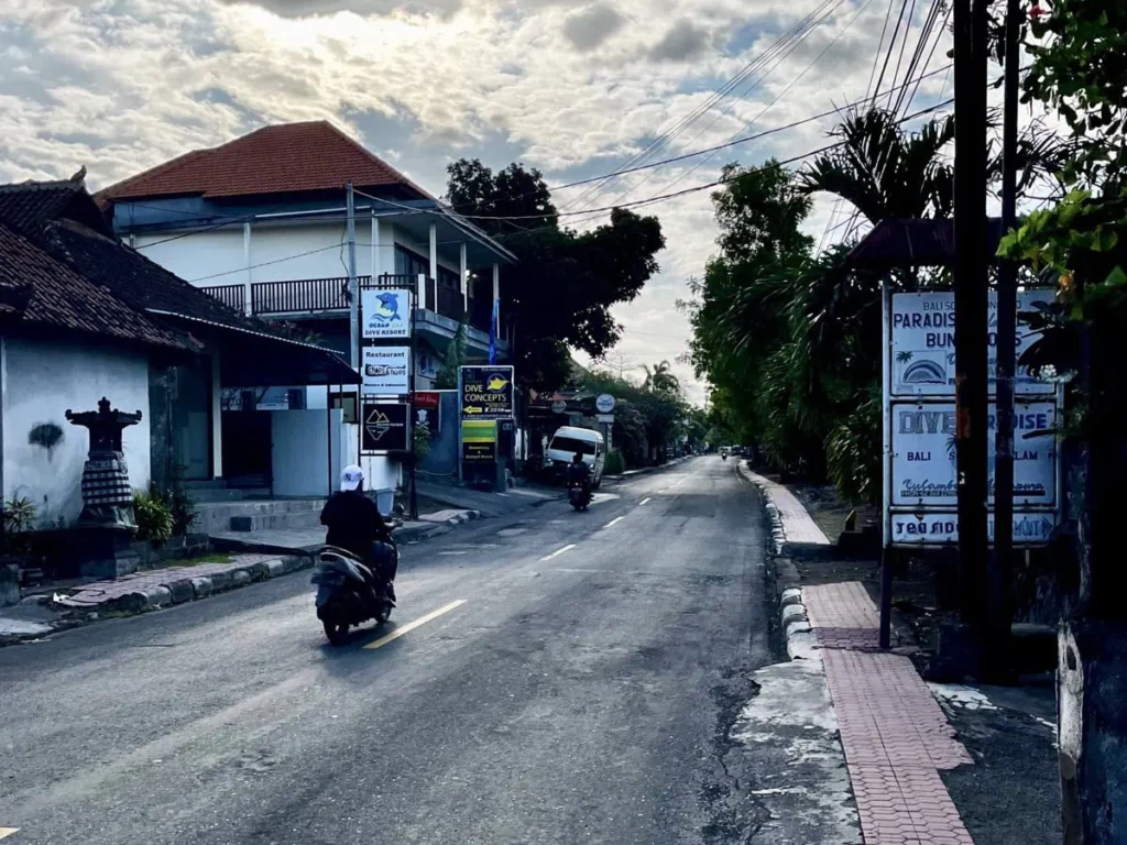 Die besten Aktivitäten in Tulamben zwischen den Tauchgängen 5 Ruhige Hauptstraße in Tulamben mit Tauchshops und kleinen lokalen Geschäften, während eine Person mit dem Scooter durch das Dorf fährt. Das Bild zeigt die typische Art, sich in Tulamben fortzubewegen, denn mit dem Scooter erreichst du Tauchplätze und Restaurants am einfachsten.