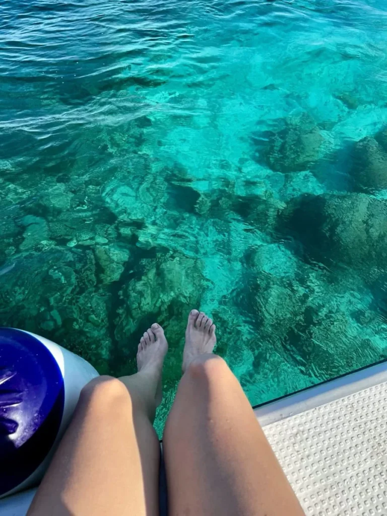 Doris sitting on the edge of a boat with their feet dangling above clear turquoise water near Semporna. Coral formations are visible beneath the surface through the calm sea.