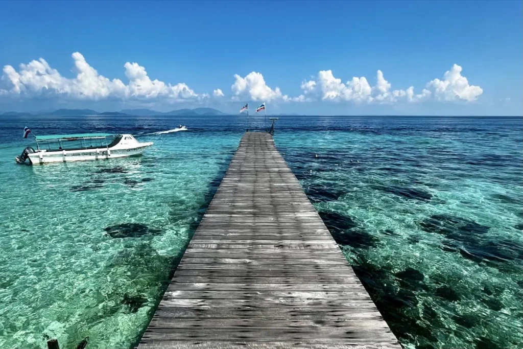 Wooden jetty stretching into clear turquoise water with a dive boat moored beside it on Mabul Island. The calm sea and bright sky mark a typical departure point for diving trips around Sipadan.