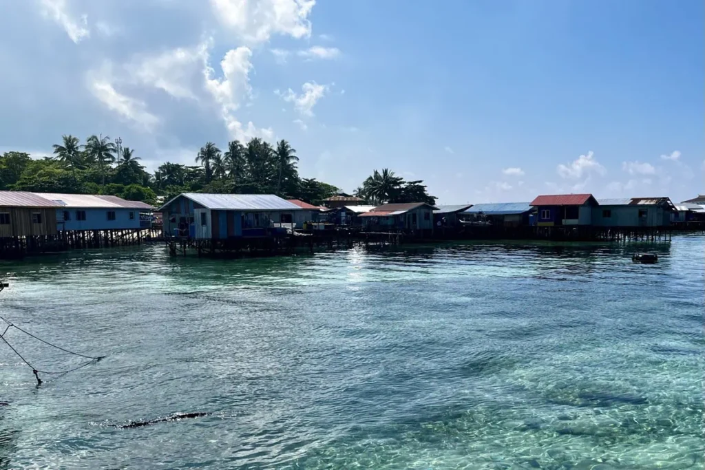 Traditional wooden houses built on stilts above shallow turquoise water in the Mabul Island water village. Palm trees and small boats line the shore of the island near Sipadan.