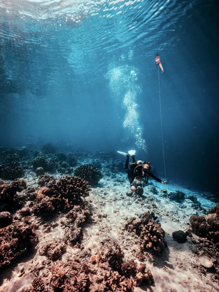 Scuba diver hovering above a shallow coral reef while performing a safety stop on a dive near Semporna. A surface marker buoy floats above in the clear blue water.