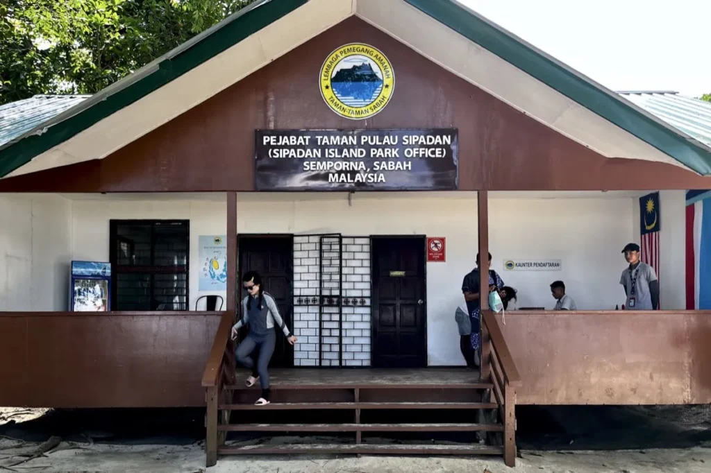 Visitors entering the Sipadan Island Park office in Semporna Sabah Malaysia with a sign reading "Pejabat Taman Pulau Sipadan Sipadan Island Park Office Semporna Sabah Malaysia" above the entrance. This is where divers check in and verify their permits before their Sipadan dive day.