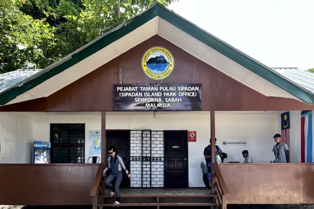 How to Get a Sipadan Permit (2026 Guide) 1 Visitors arrive at the Sipadan Island Park Office in Semporna Malaysia where you obtain a Sipadan permit. A sign reads "Pejabat Taman Pulau Sipadan (Sipadan Island Park Office) Semporna Sabah Malaysia" above the entrance as staff assist at a registration counter.