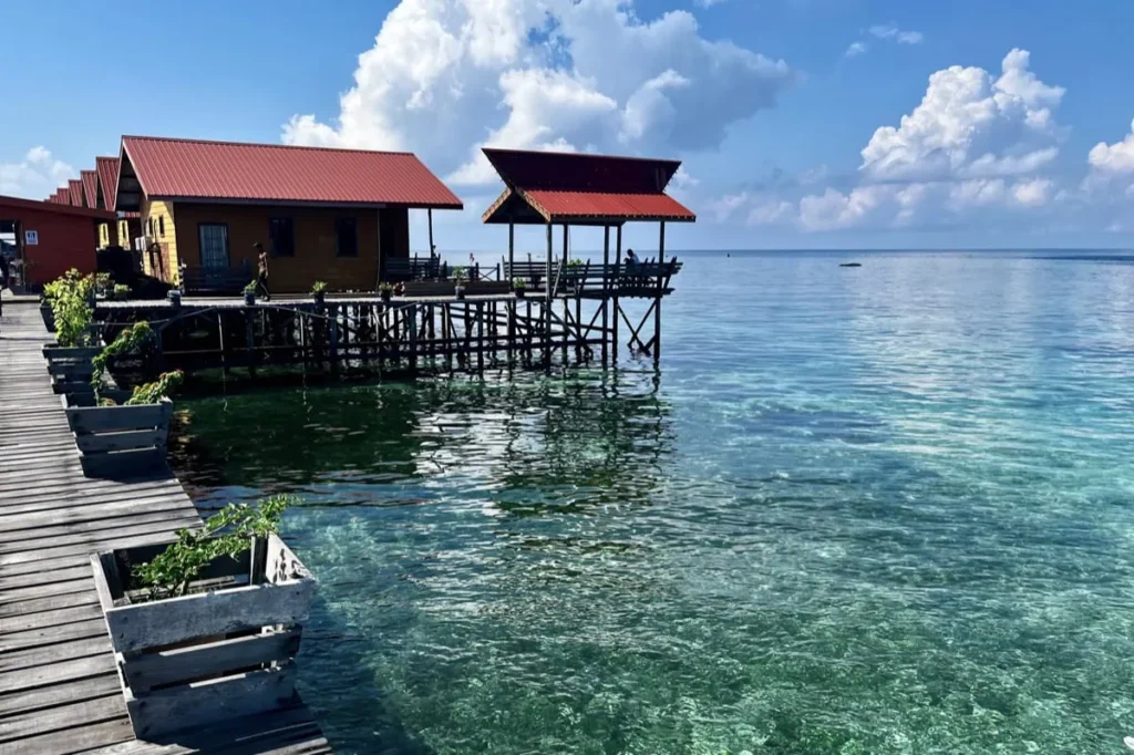 Wooden stilt buildings of Uncle Chang Dive Resort standing above shallow turquoise water on Mabul Island. A long boardwalk connects the huts over the sea.