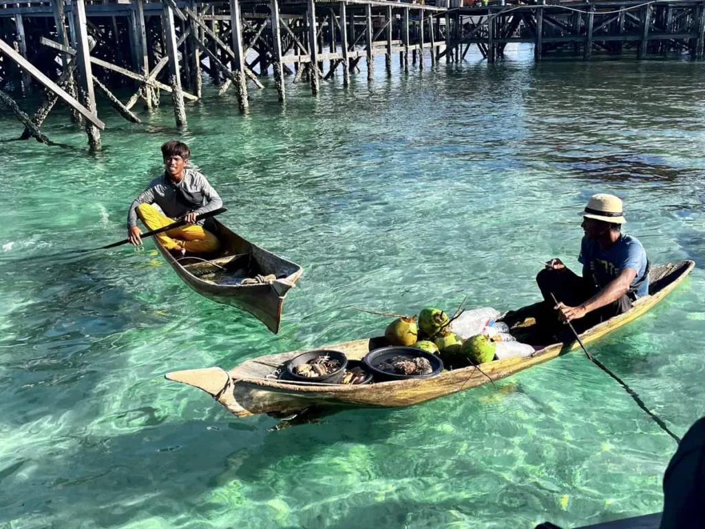 Two Bajau Laut men paddle wooden boats in shallow clear water at Mabul Island with fresh coconuts and seafood, offering a glimpse of local life near Sipadan dive resorts.