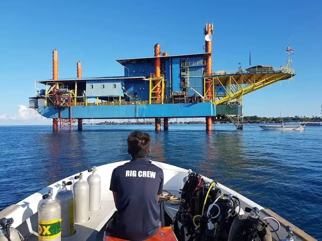 Diver on a boat approaches the Seaventures dive rig, a converted offshore platform used as unique accommodation near Sipadan with tanks and gear visible on deck.