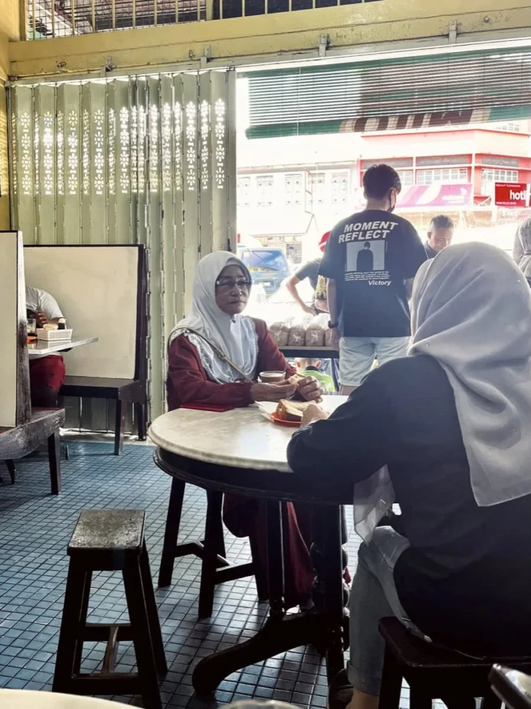 Two women wearing headscarves sit at a small round table inside a casual cafe in Semporna with simple tiled floors and open shutters to the street.