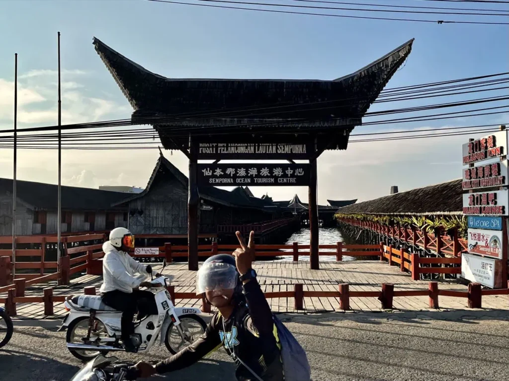 Entrance to Semporna Ocean Tourism Centre and Semporna jetty with a wooden walkway over water and a large traditional roofed gate while motorbikes pass by in the foreground.