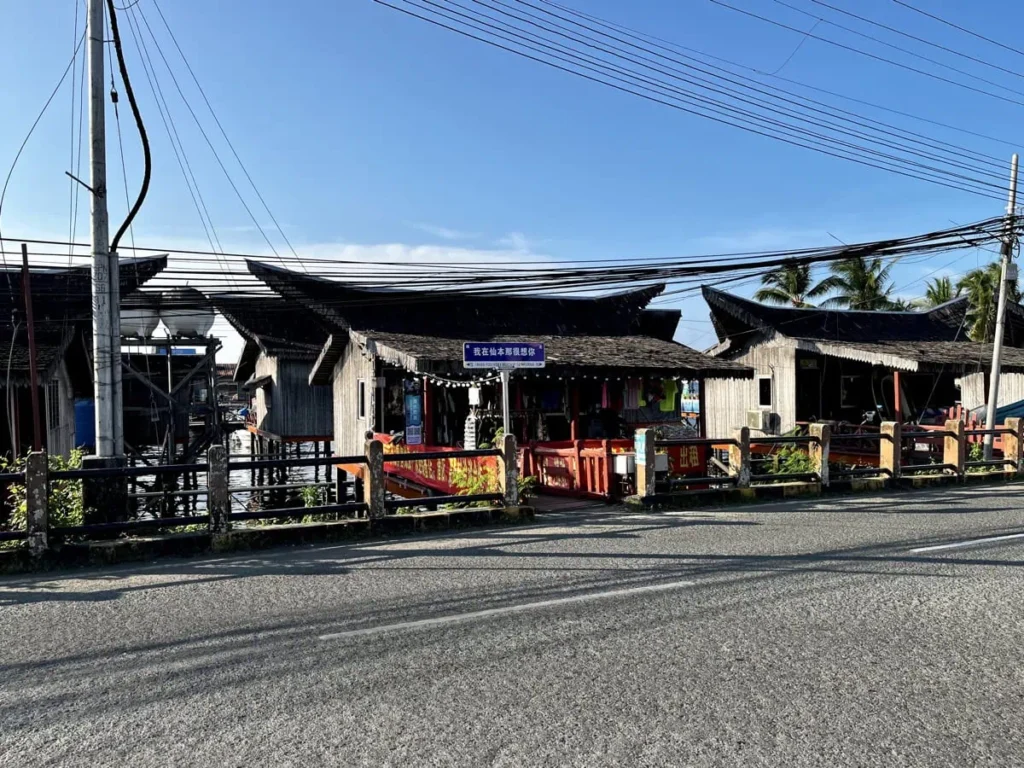 Wooden stilt houses and small shops line a roadside in Semporna with power lines overhead and a glimpse of water behind, showing the town where travelers stay before visiting Sipadan.